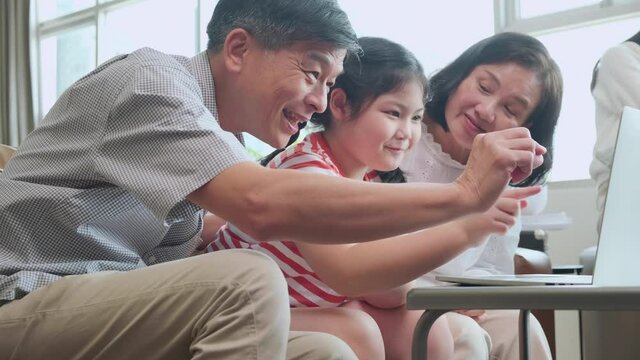 grand mother grandfather teach little child granddaughter using laptop sit on sofa, happy relation bonding generation asian family spend time online playing with laptop together at home