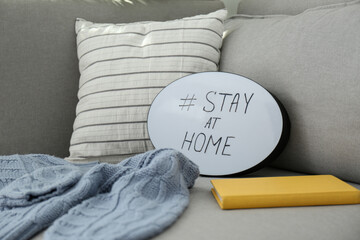 Book, sweater and speech bubble with hashtag STAY AT HOME on sofa, closeup. Message to promote self-isolation during COVID‑19 pandemic