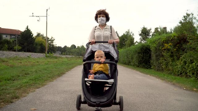 A grandmother with a medical mask pushing an upset baby boy in a stroller on a street. Covid-19 separating families.