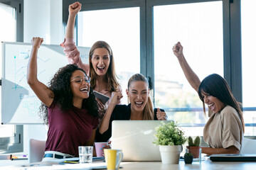 Four happy smart business women work with laptops on the desk while celebrating a victory in the...