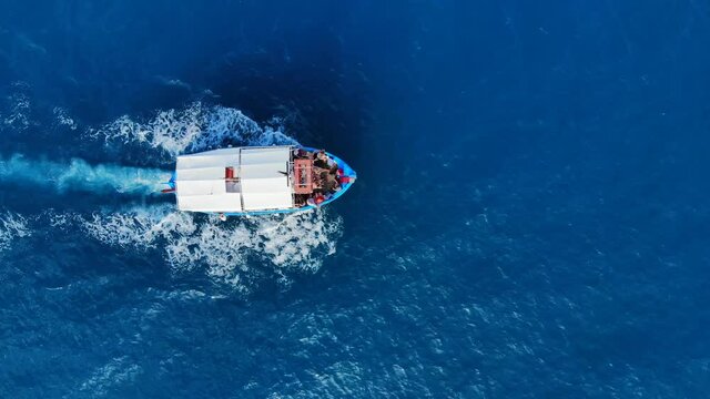 Short, wide boat slowly sail at clear sea, top-down aerial shot. Small tourist watercraft with people on board, return to port after trip. Beautiful blue colour of crystal water at Aegean sea