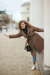 A portrait of a curly-haired young woman peeking out from behind a white pillar. The girl laughs and stretched out her hand to the side, catching the car