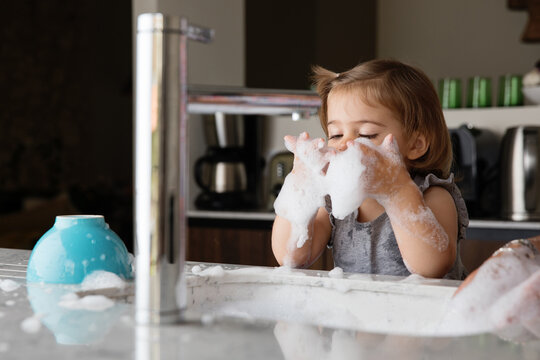 Cute toddler girl playing with suds while wahsing dishes in kitchen sink