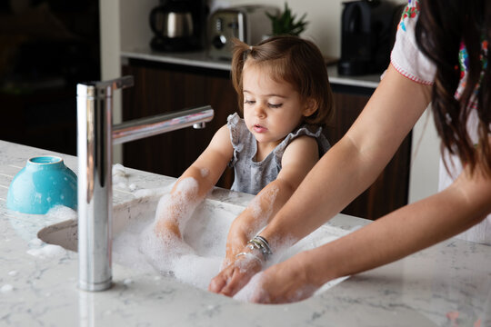 Cute toddler girl helping mother wahsing dishes in kitchen sink