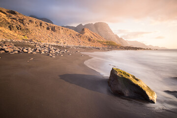Atardecer en la costa montañosa. Playa con rocas y olas que van alisando la arena. El sol del atardecer da volumen y profundidad.