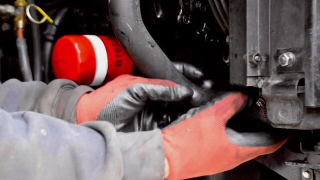 Adult hands with red and black protective gloves drains antifreeze out of diesel generator by unscrewing screw with driver and pulling pipe as red liquid exits, static close up profile