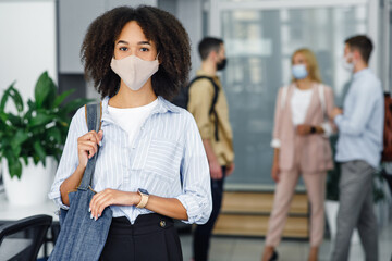 Social distancing and office work with colleagues. Attractive young african american woman with bag in protective mask in coworking office interior during virus outbreak