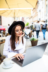 Smiling young woman with laptop sitting in street cafe, browsing web and social network feed, texting with friends