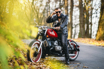 Fototapeta na wymiar Bearded brutal man in sunglasses and leather jacket sitting on a motorcycle on the road in the forest