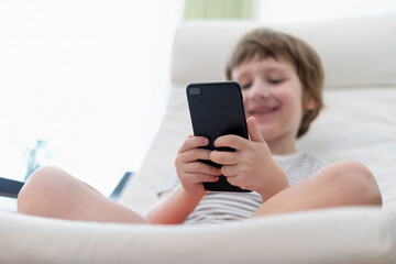 Little child boy playing online game, watching video on smartphone, sitting on couch entertaining in living room. Small kid using funny mobile apps, enjoying free leisure time at home