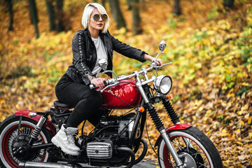 Fototapeta na wymiar Pretty blonde biker girl in sunglasses with red motorcycle on the road in the forest