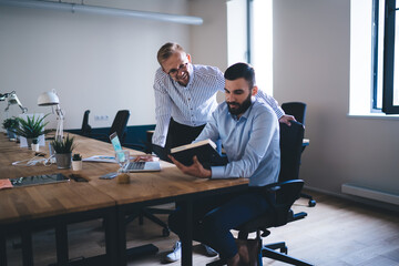 Smiling male employees sitting at table together reading report and analyzing accountings for startup enjoying cooperation, professional men designers talking to each other and having discussion