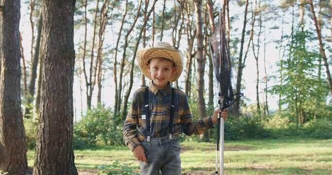 Handsome smiling small fishman in straw hat posing on camera with fishing rod and landing net in the middle of green park,slow motion