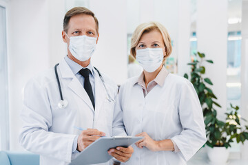 Adult female and male doctors standing together on clinic foyer while looking at camera wearing face masks for prevention epidemic.