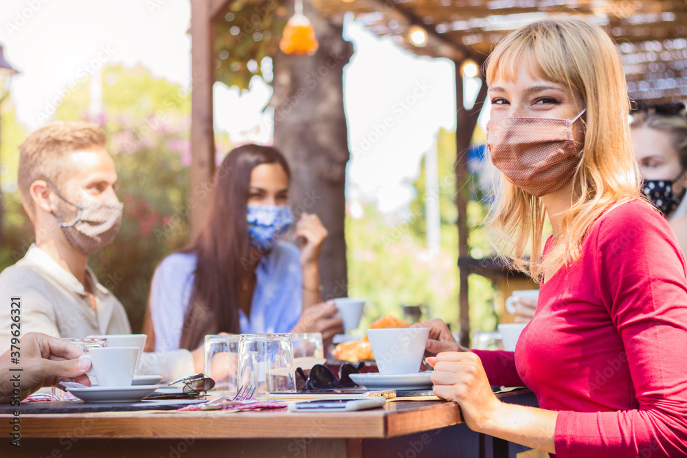 Wall mural Happy young woman wearing face mask smiling at the camera at the restaurant cafè. Group of friends drinking coffee sitting at the bar. New normal concept.