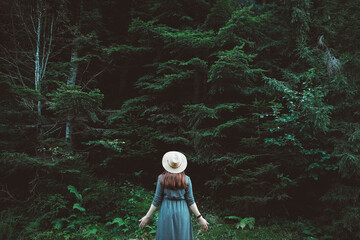 Stylish young woman in a straw hat and dress stands on a background of green forest and trees. Travel concept. Copy, empty space for text
