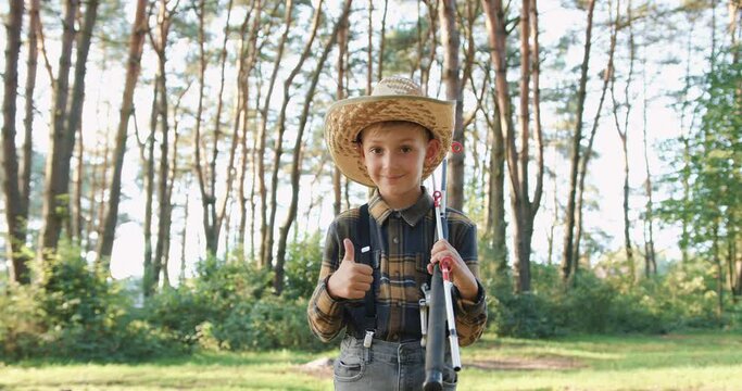 Front view of good-natured satisfied smiling happy 10-aged boy in straw hat which posing on camera with fishing rods in the green park and showing sign ok