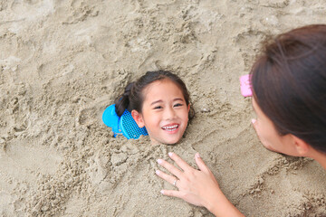 Happy asian little child girl buried in the sand at the beach by mother. Kid playing with sand at...