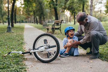 boy fall from his bike in the park. grandfather helping him