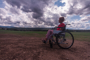 wide angle disabled girl sitting in a wheelchair in the middle of a green field in the countryside with a sky clouds