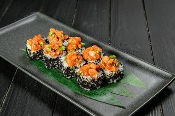 Side view of Japanese sushi volcano roll with black flying fish roe (tobiko). Marinated salmon and spring onions on top. Served on bamboo leaves on black plate. Dark wooden background
