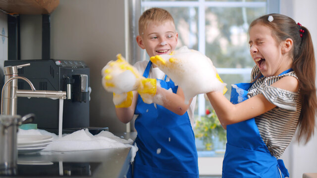 Happy Boy And Girl Kids Washing Dishes And Playing With Foam In Kitchen