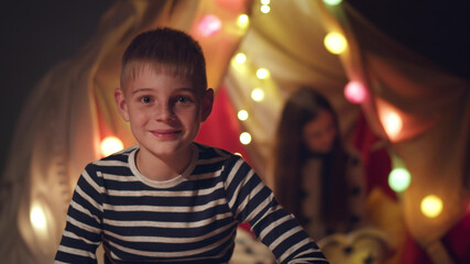 Happy smiling preteen boy in pajama smiling at camera with sister sitting in wigwam on background © nimito