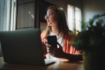 Young pensive millennial girl in glasses sitting at workplace near window holding smartphone and...