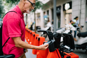 Mature male tourist using mobile application for making contact payment and rent electronic bicycle for city sightseeing on way, aged Caucasian man connecting to banking app for web pay on street
