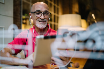 Caucasian senior in classic spectacles and electronic headphones using media application on modern touch pad for watching online video, man in earphones listening music while making web booking