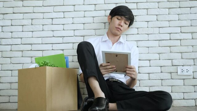 Business & unemployment concept. Low angle shot of desperate young Asian man sitting near the packed stuff in the box for moving out, leans on the white bricks wall, looking at picture frame sadly