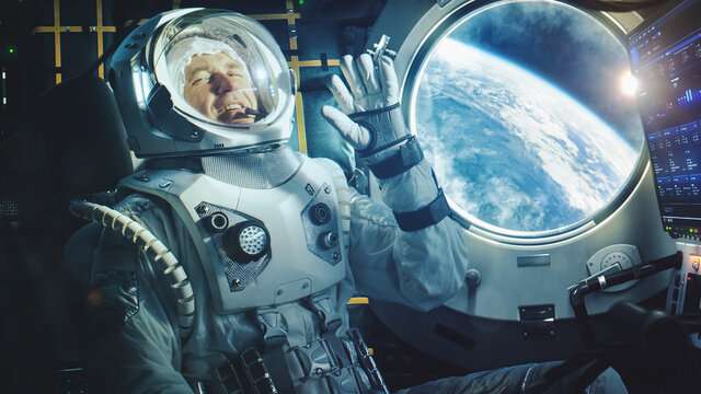Portrait of a Happy Astronaut on a Space Ship In Orbit. Cosmonaut in a Futuristic Space Suit is Full of Joy and is Waving Hand on a Video Call. VFX Graphics Shot from the International Space Station.