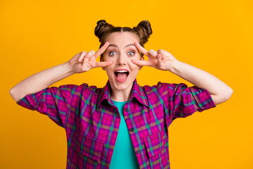 Photo of attractive excited funky teen lady two funny buns good mood overjoyed showing v-sign symbol open mouth see friends wear casual plaid shirt isolated yellow bright color background