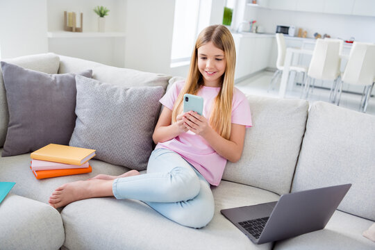 Full size photo of positive little kid girl sit cozy couch study remote use smartphone have social media online classmates communication in house indoors