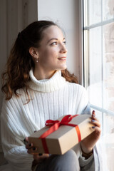 Beautiful young woman with a gift box sitting near window at home and smiling. Cozy winter and autumn time.