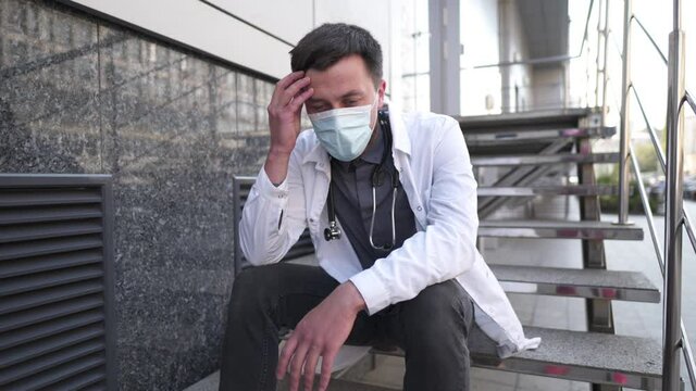 Worried and tired doctor sitting on stairs outside hospital. Exhausted physician in white lab coat and medical protective mask, sit on staircase clinic. Stress healthcare worker during COVID-19.