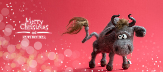 Christmas banner. The bull is a symbol of the New year 2021. Knitted brown toy bull on a red background with bokeh and the inscription happy Christmas and New year. Close-up, selective focus.