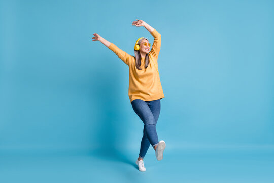 Full body size photo of attractive careless old dancer lady listen music earphones hands waving above head walking street wear sunglass yellow jumper jeans vivid blue color background
