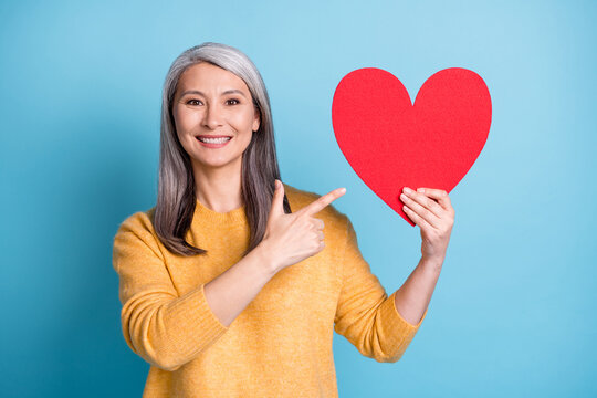 Perfect valentine present. Photo of cute attractive mature lady white grey hairdo hands hold direct finger paper red heart figure wear yellow sweater isolated blue pastel color background