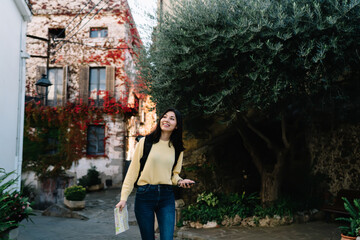 Cheerful female backpacker in casual clothing enjoying travel sightseeing around touristic village, carefree woman with location map and smartphone walking around town street during solo journey