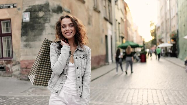 Beautiful fashionable model with curly hair holding shopping bags on her shoulder, looking at the camera and smiling. Portrait of young beautiful woman. Female nature beauty. Slow motion