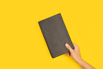 Mano femenina sosteniendo un libro de tapa negro sobre un fondo amarillo liso y aislado. Vista de...