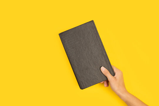 Mano femenina sosteniendo un libro de tapa negro sobre un fondo amarillo liso y aislado. Vista de frente. Copy space