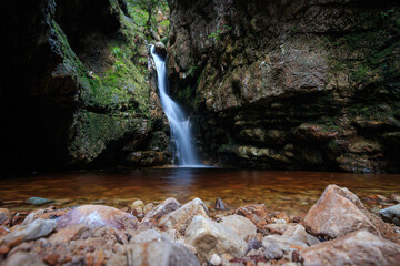 Waterfall between mossy cliffs pouring into rocky pool in a dark mountain gorge. Western Cape, South Africa.