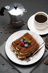 portion of Classic tiramisu dessert with raspberries and blueberries, cup of espresso and coffee maker on dark concrete background