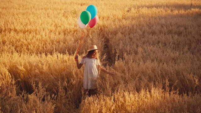 girl in white dress and straw hat walking with colorful balloons in wheat field
