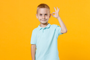 Smiling handsome little male fair-haired brown-eyed kid boy 5-6 years old wearing stylish blue turquoise t-shirt polo showing ok gesture isolated on yellow color wall background, child studio portrait