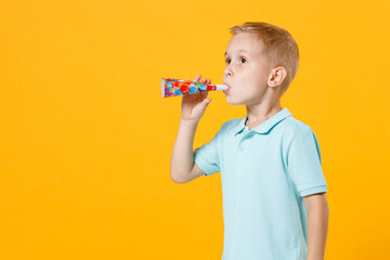 Little cute fun male kid boy 5-6 years old wearing stylish blue turquoise t-shirt polo celebrate hold in hands and blow in birthday pipe isolated on yellow color wall background, child studio portrait