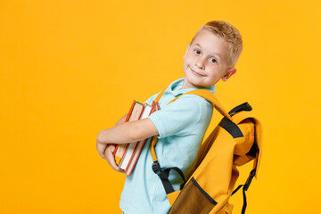 Fototapeta Little fun male kid boy 5-6 years old wearing stylish blue turquoise t-shirt polo backpack holding in hands school books isolated on yellow color background, child studio portrait. Education concept. obraz
