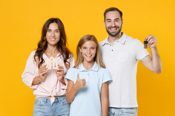 Smiling young parents mom dad with child kid daughter teen girl in basic t-shirts hold house bunch of apartment keys showing thumb up isolated on yellow background studio portrait. Family day concept.